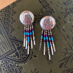 May include: A pair of circular silver earrings featuring an engraved design of a wolf howling at a moon. Each earring has five dangling strands composed of alternating small silver beads, purple cylindrical beads, and bright blue cylindrical beads.