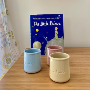 May include: Three ceramic pots in light blue, soft pink, and pale yellow, each embossed with the text la fermière, positioned in front of a blue hardcover book titled The Little Prince by Antoine de Saint-Exupéry featuring a yellow star illustration.