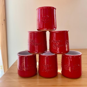 May include: A stack of six red ceramic jars. Each jar has a cylindrical shape with a slightly flared rim. Some jars feature embossed lips, while others have the phrases "Be Mine" or "Bisou Bisou".