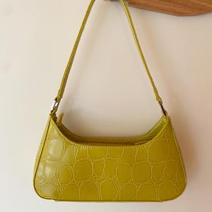 Puede incluir: Un bolso baguette verde lima con textura de piel de cocodrilo. El bolso tiene forma curva, cierre de cremallera y una fina correa para el hombro. El bolso est&aacute; colgado de un gancho de pared de madera.