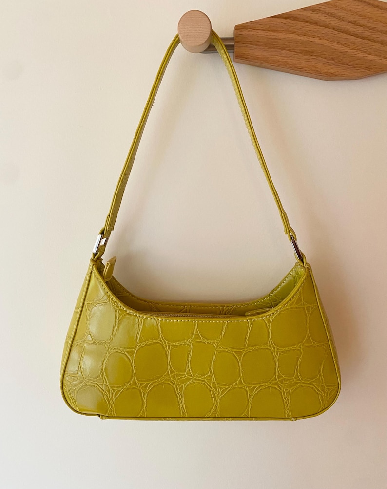 Puede incluir: Un bolso baguette verde lima con textura de piel de cocodrilo. El bolso tiene forma curva, cierre de cremallera y una fina correa para el hombro. El bolso est&aacute; colgado de un gancho de pared de madera.
