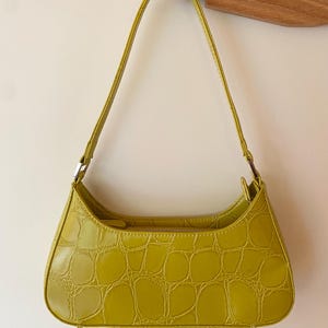Puede incluir: Un bolso de hombro verde lima con textura de piel de cocodrilo. El bolso tiene forma curva, cierre de cremallera y una correa fina. El bolso est&aacute; colgado de un gancho de madera.