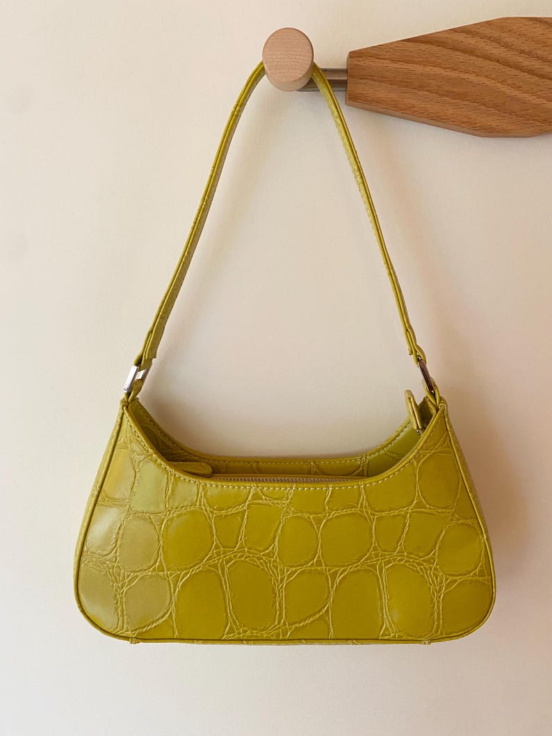 Puede incluir: Un bolso de hombro verde lima con textura de piel de cocodrilo. El bolso tiene forma curva, cierre de cremallera y una correa fina. El bolso est&aacute; colgado de un gancho de madera.