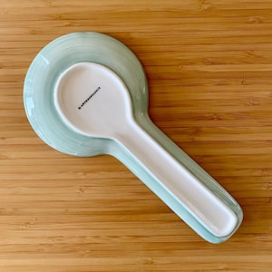May include: A white ceramic spoon and spoon rest set. The spoon rest is a light green color with a matching spoon. The spoon rest has a circular base and a long, narrow trough for the spoon. The spoon has the text "by ANTHROPOLOGIE".