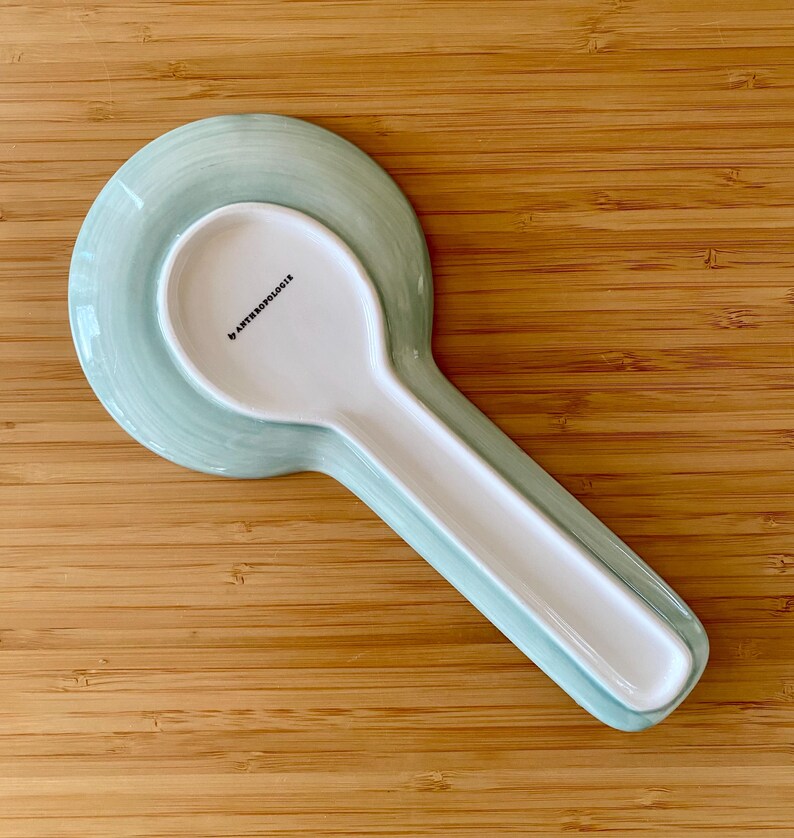 May include: A white ceramic spoon and spoon rest set. The spoon rest is a light green color with a matching spoon. The spoon rest has a circular base and a long, narrow trough for the spoon. The spoon has the text "by ANTHROPOLOGIE".