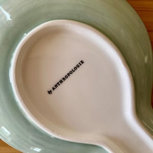 May include: A ceramic spoon rest with a light green, watercolor-style design. The rest has a wide, oval base and a long, narrow handle. The words "by ANTHROPOLOGIE" are printed in black on the white interior.