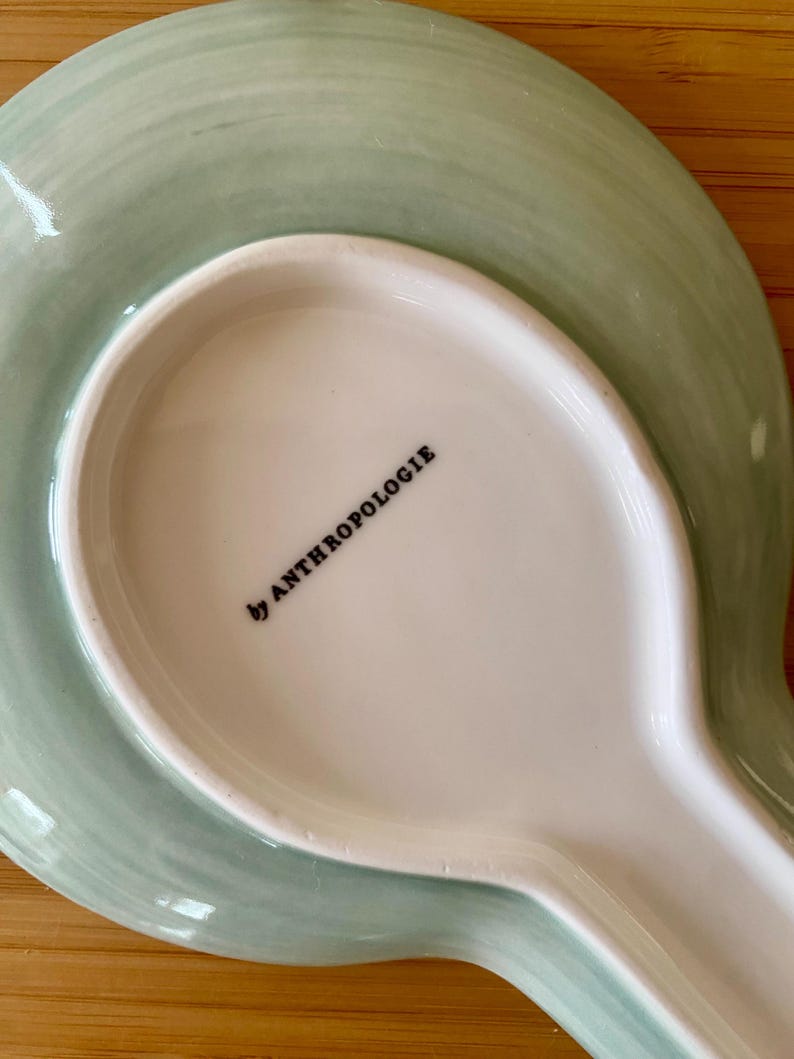 May include: A ceramic spoon rest with a light green, watercolor-style design. The rest has a wide, oval base and a long, narrow handle. The words "by ANTHROPOLOGIE" are printed in black on the white interior.