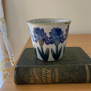 May include: A ceramic planter with a white base and a hand-painted pattern of blue flowers and green leaves. The planter is resting on a dark green hardcover book titled Mrs. Partington's Knitting Work by Shillaber, published by Lee and Shepard.