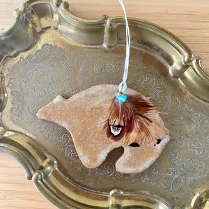 May include: A tan-colored bear-shaped ornament with a cluster of brown and black feathers and a small turquoise bead. The ornament hangs from a white string and rests on a gold-colored tray with ornate detailing.