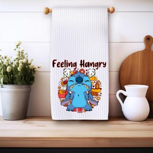 May include: White kitchen towel with a blue cartoon character with the text "Feeling Hangry" printed on it. The character is surrounded by images of food.