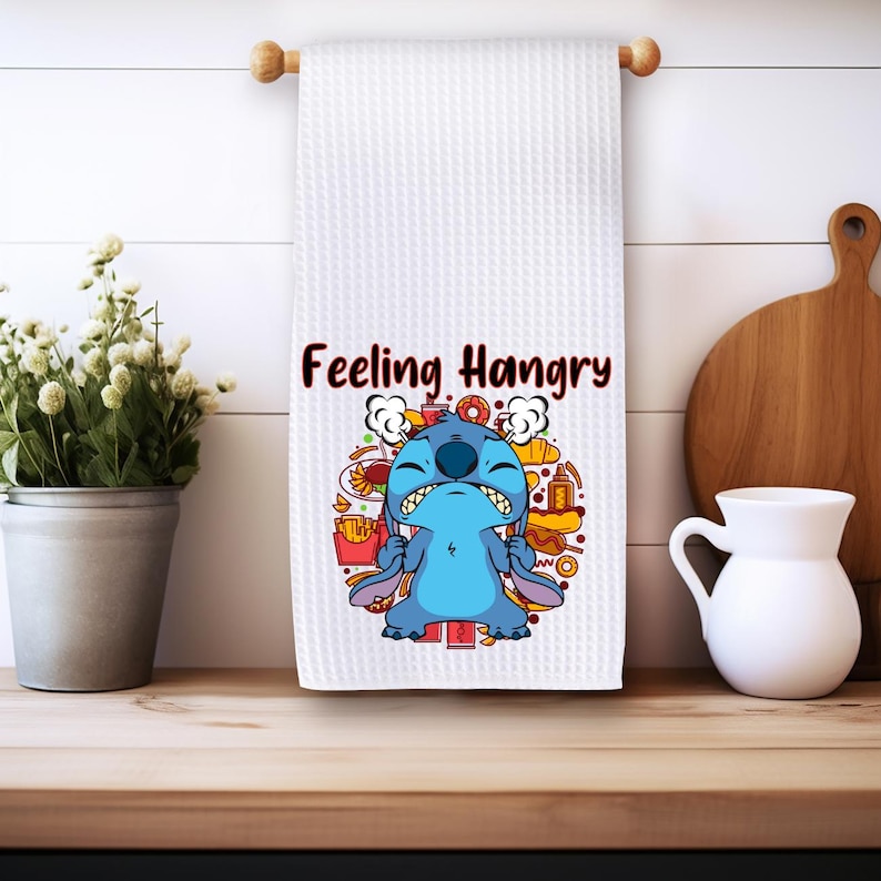 May include: White kitchen towel with a blue cartoon character with the text "Feeling Hangry" printed on it. The character is surrounded by images of food.