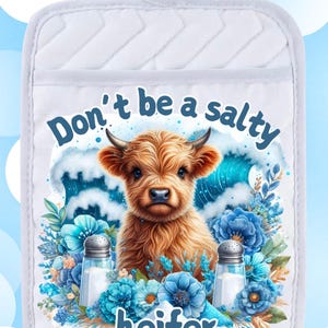Puede incluir: Un agarrador de olla blanco con una linda ilustración de una vaca de las tierras altas y el texto "Don't be a salty heifer". La vaca está rodeada de flores azules y saleros.