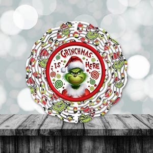 May include: A white and red circular wind spinner with a green Grinch face in the center. The text "Grinchmas is here" is written in a red circle around the Grinch. The background of the spinner is white with red and green candy cane stripes.
