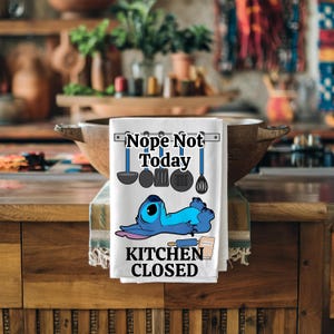 Pode incluir: Toalha de cozinha branca com o texto "Nope Not Today" e "Kitchen Closed". A toalha apresenta uma personagem de desenho animado azul deitada com um rolo. Utensílios de cozinha são retratados acima da personagem.