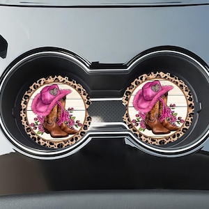 May include: Two pink and brown car coasters with a leopard print border. Each coaster features a pink cowboy hat and brown cowboy boots on a white wood background.