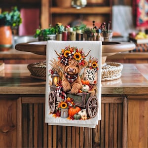 May include: A white kitchen towel featuring a graphic of a fluffy highland cow wearing a plaid scarf and flower crown, sitting in a wooden wagon with pumpkins, sunflowers, a lantern, and a sign that reads Hello Fall in black script.