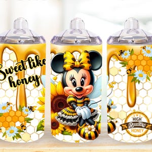 May include: A white tumbler with a clear lid and handle. The tumbler features a design of a cartoon mouse dressed as a bee with a honeycomb background. The text "Sweet like honey" is printed on the tumbler.