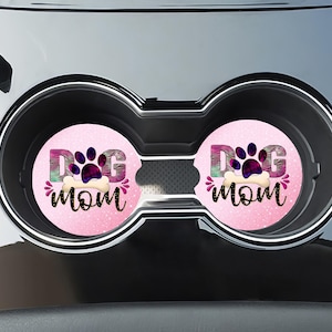 May include: Two pink car coasters with a white bone and paw print design. The coasters say "Dog Mom" in a pink and purple font.