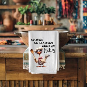 May include: White kitchen towel with the text "GO AHEAD SAY SOMETHING ABOUT MY Cooking". The towel features a cartoon chicken wearing a chef's hat and holding a wooden spoon. The chicken is wearing a striped apron and a red bow tie.