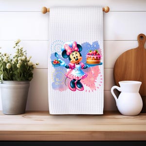 May include: White waffle weave kitchen towel with a colorful illustration of Minnie Mouse holding a cake and a plate of strawberries. The background is a watercolor wash of pink and blue with white fireworks.
