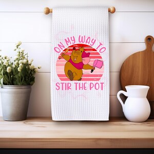 Puede incluir: Una toalla de cocina blanca con un gráfico de puesta de sol rosa y rojo. El gráfico presenta un ratón de dibujos animados sosteniendo una cuchara y una olla. El texto "On My Way To Stir The Pot" está escrito en letras rosas.