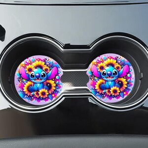 Puede incluir: Dos posavasos para coche con un personaje de dibujos animados azul con orejas grandes, rodeado de girasoles. Los posavasos están colocados dentro de los portavasos de un coche. El diseño incluye colores rosa, amarillo, morado y azul.