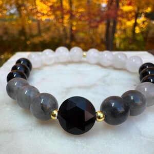 May include: A beaded bracelet featuring a large, faceted black bead as a focal point, surrounded by gray, black, and white spherical beads. Gold-colored spacers are interspersed throughout the design. The bracelet is displayed on a white surface.