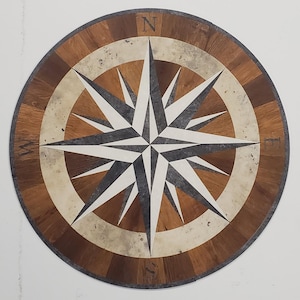 May include: Circular wooden compass rose inlay with a starburst design. The compass rose features a central star with alternating gray and white points, surrounded by a light beige and dark brown wood grain pattern. The cardinal directions are subtly marked.