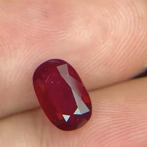 Könnte beinhalten: Ein tiefroter, oval geschliffener Edelstein mit einer facettierten Oberfläche, die das Licht reflektiert. Der Stein zeigt einen satten, kräftigen karmesinroten Farbton und eine polierte Oberfläche mit mehreren geometrischen Facetten auf der Oberseite.