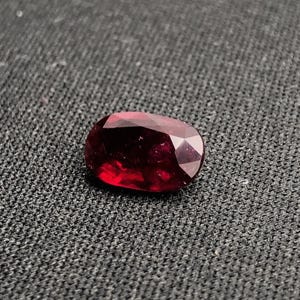 May include: A deep red, oval-cut gemstone with multiple facets. The polished surface of the gem reflects light, revealing its internal structure. The stone is set against a dark, textured background, accentuating its colour and clarity.