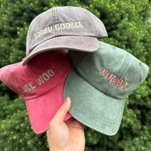 May include: Three baseball caps in washed-out colours. The top cap is grey with the embroidered text GOOFY GOOBER. The bottom left cap is red with the embroidered text WEE WOO. The bottom right cap is sage green with the embroidered text WUMBO.