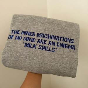 May include: A heather gray sweatshirt featuring dark blue embroidered text that reads: THE INNER MACHINATIONS OF MY MIND ARE AN ENIGMA *MILK SPILLS*. The garment is folded and displays a textured fabric surface with clear, bold lettering.