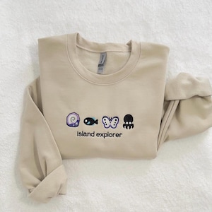 May include: A beige sweatshirt with a graphic of a seashell, fish, butterfly, and octopus embroidered on the front. The text "Island explorer" is embroidered below the graphic.