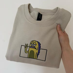 May include: A beige crewneck sweatshirt featuring an embroidered graphic of a yellow fish character with purple fins, making a peace sign gesture. The character is enclosed within a thin black rectangular outline on the center of the chest.