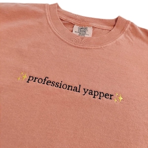 May include: A peach-coloured t-shirt with the text "professional yapper" embroidered in black with gold glitter stars on either side.