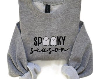 May include: A heather gray sweatshirt with the words "SP00KY season" in black script and two ghost illustrations. The sweatshirt has a crew neck and long sleeves with gray cuffs.