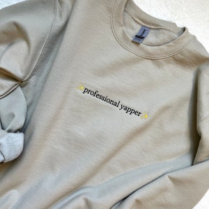 May include: A light beige sweatshirt with the text "professional yapper" embroidered in black with yellow stars on either side.