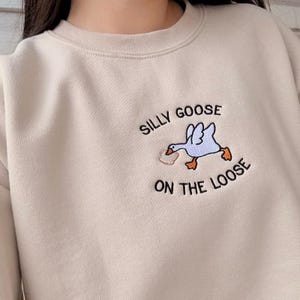 May include: A beige sweatshirt with a black embroidered design of a goose carrying a piece of bread with the text "Silly Goose on the Loose".