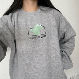 May include: A gray sweatshirt featuring a green lizard graphic inside a black rectangle. The text "LIZARD LIZARD LIZARD" is printed below the graphic. The sweatshirt is made of a soft material.