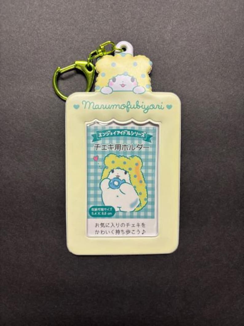 May include: A light yellow Marumofubiyori keychain photo holder with a cartoon character design. The holder has a rectangular shape with a clear window for a photo and a green clasp. The text "Marumofubiyori" is visible.