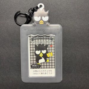 May include: A gray Badtz-maru photo holder with a black keyring. The holder features a cartoon Badtz-maru character and text in Japanese. The holder is designed to hold a photo that is 5.4 x 8.6 cm.