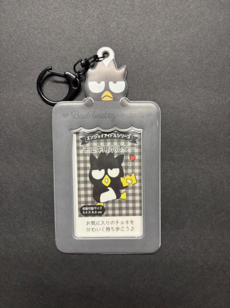 May include: A gray Badtz-maru photo holder with a black keyring. The holder features a cartoon Badtz-maru character and text in Japanese. The holder is designed to hold a photo that is 5.4 x 8.6 cm.