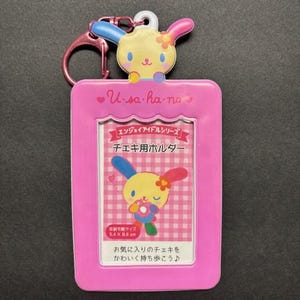 May include: A pink Usahana ID holder with a cartoon bunny design. The holder has a pink keychain and a clear window for displaying a card. The text "U-sa-ha-no" is printed on the top.