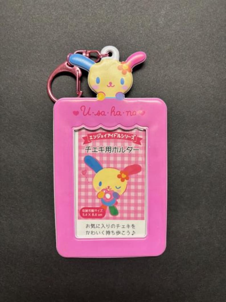 May include: A pink Usahana ID holder with a cartoon bunny design. The holder has a pink keychain and a clear window for displaying a card. The text "U-sa-ha-no" is printed on the top.