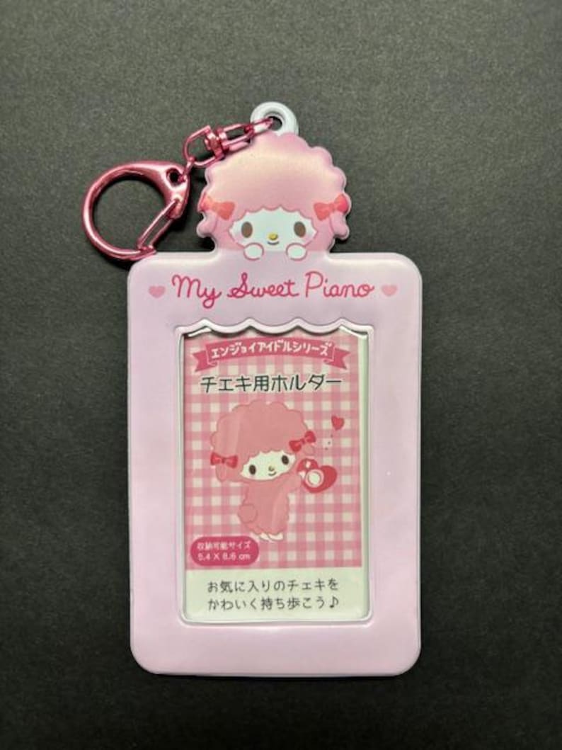 May include: A pink photo holder keychain featuring a cartoon sheep character with a pink bow. The holder has a rectangular frame with the text "My Sweet Piano" and Japanese text. The keychain includes a pink clasp.