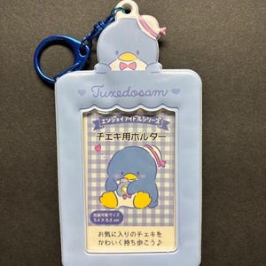 May include: A light blue Tuxedosam photo holder with a blue keyring. The holder features a cartoon penguin and text in Japanese. The photo holder is rectangular with rounded edges.