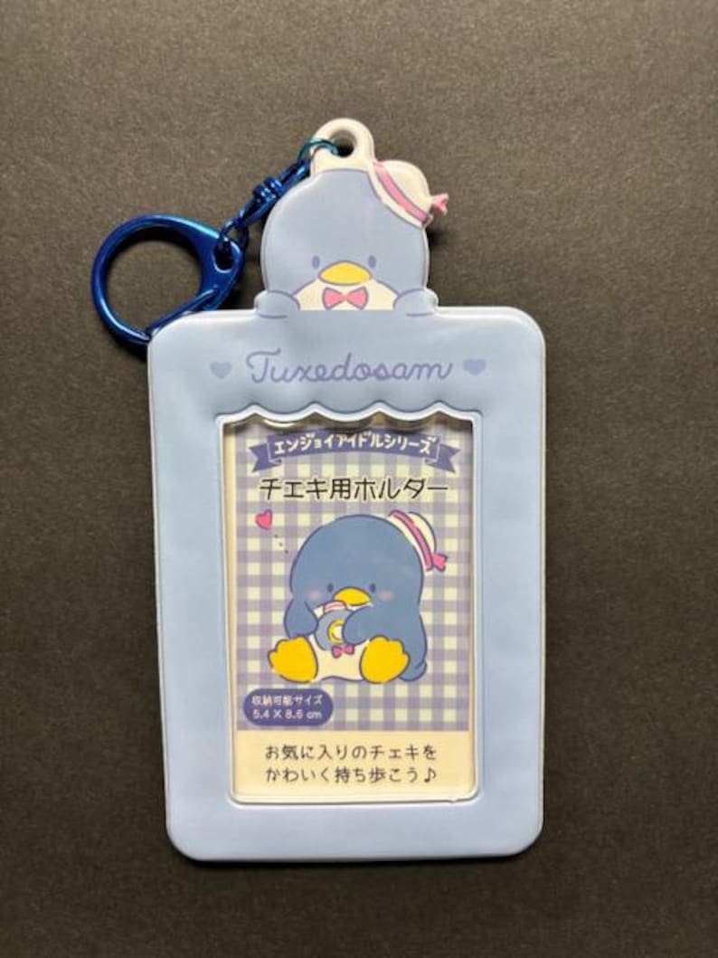 May include: A light blue Tuxedosam photo holder with a blue keyring. The holder features a cartoon penguin and text in Japanese. The photo holder is rectangular with rounded edges.