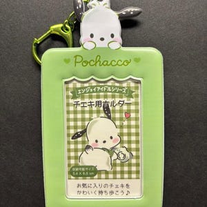 May include: A green Pochacco keychain with a cartoon image of Pochacco. The keychain has a green frame with the word "Pochacco" and Japanese text. The frame holds a picture of Pochacco with a camera. The keychain is 5.4 x 8.6 cm.