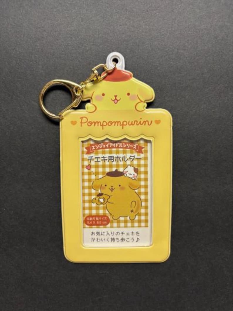 May include: Yellow Pompompurin photo holder with a gold keychain. The holder features a cartoon character design and Japanese text. The holder is designed to hold a photo that is 5.4 x 8.5 cm.