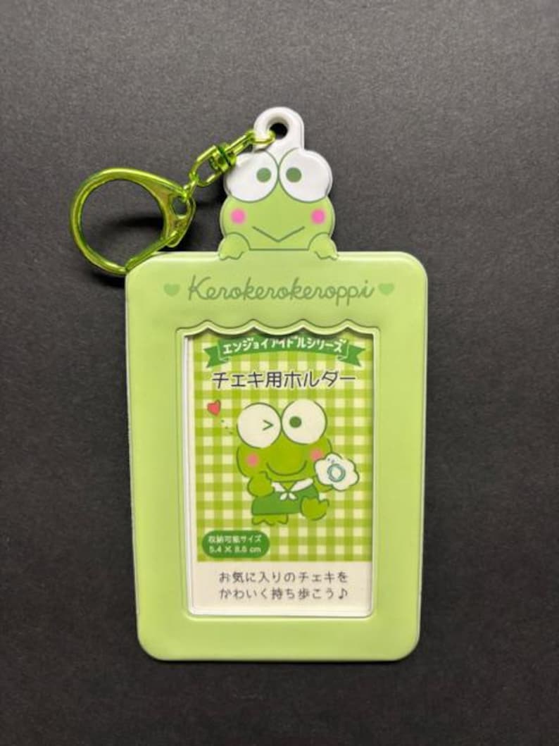 May include: A light green Keroppi photo holder with a matching green keyring. The holder features a Keroppi frog design and Japanese text. The holder is designed to hold photos that are 5.4 x 8.6 cm.